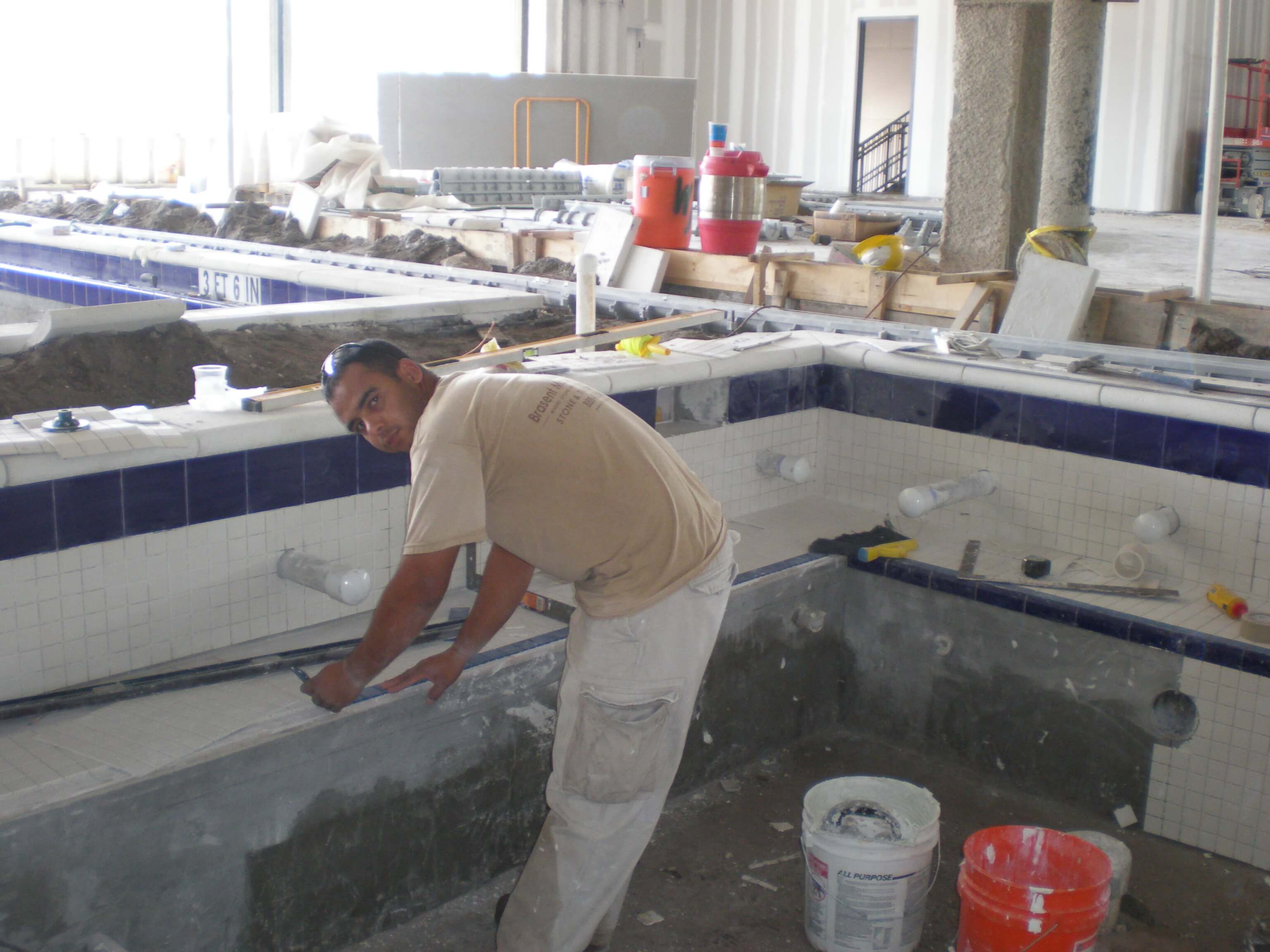 Tile installer measuring and preparing to place tiles inside a partially completed swimming pool with blue and white tiles.
