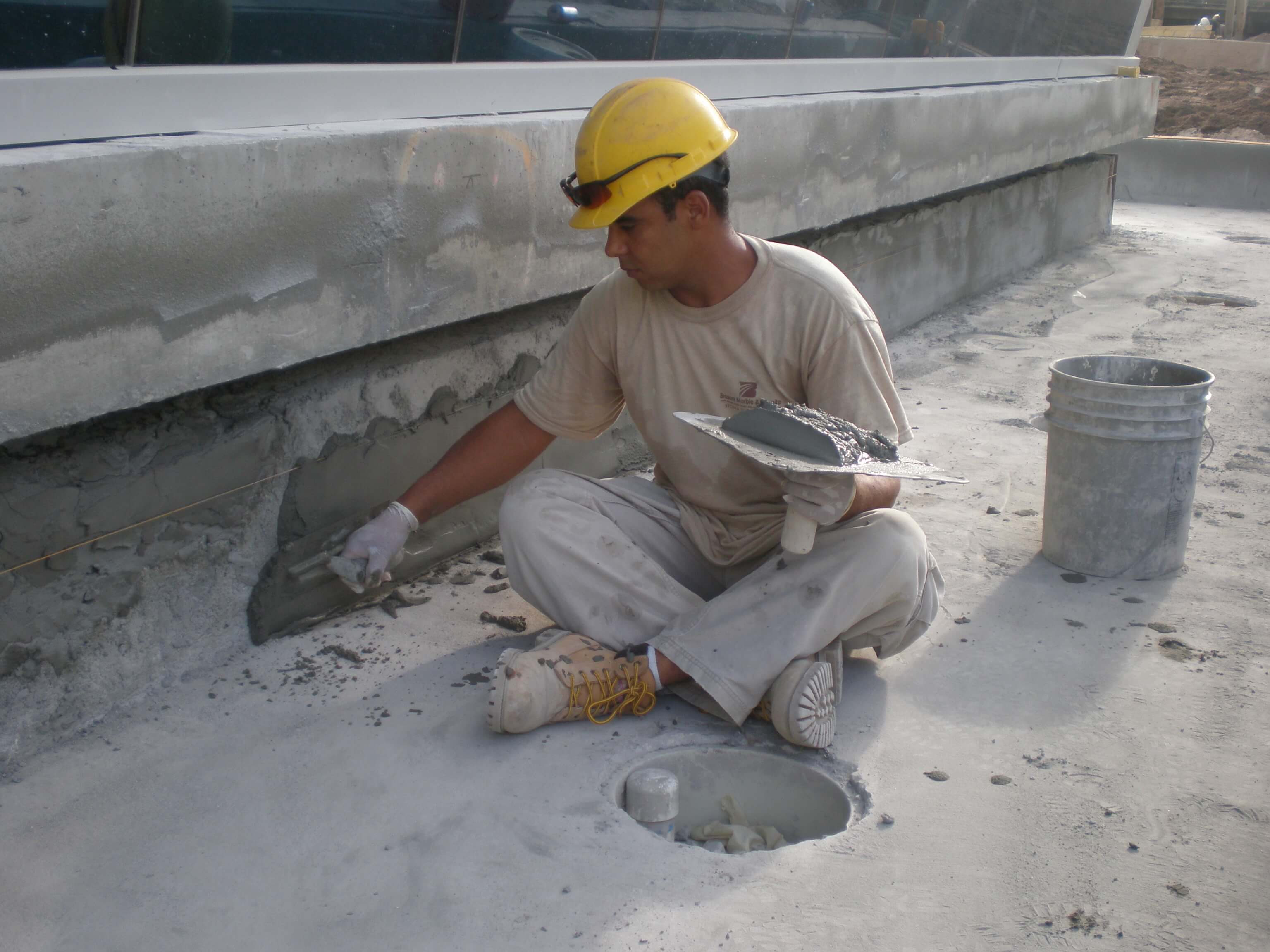 Tile installer wearing a hard hat, sitting on the ground and carefully applying mortar to a concrete surface, preparing the area for tile installation.