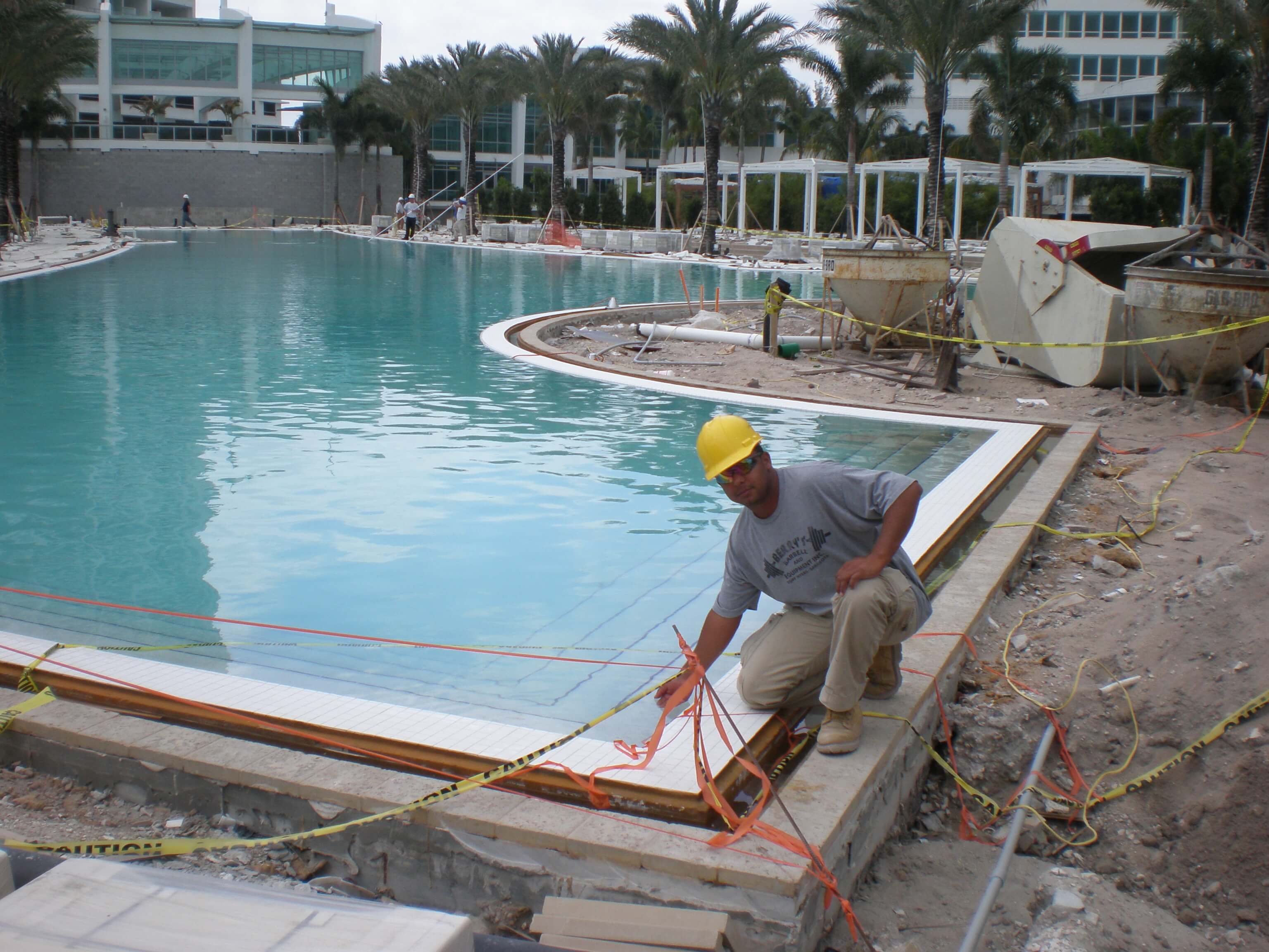 Tile installer working near the edge of a pool under construction at the Fontainebleau Miami Beach resort, with palm trees and hotel buildings in the background.